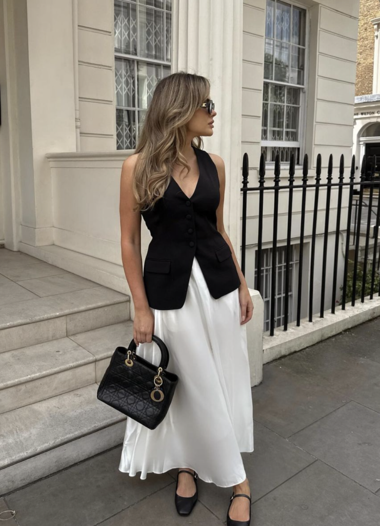 Woman standing outside wearing a black sleeveless top and white satin midi skirt, holding a designer handbag.